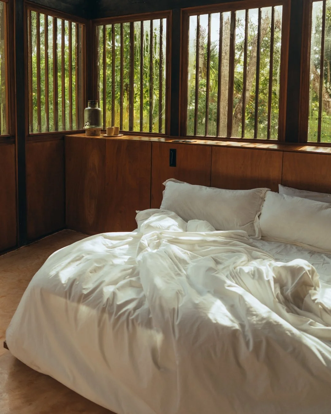 A cozy bedroom with a large bed covered in white rumpled sheets and pillows, big wooden windows overlooking a green, leafy outdoor scene, a glass jar, and cups on a wooden ledge.