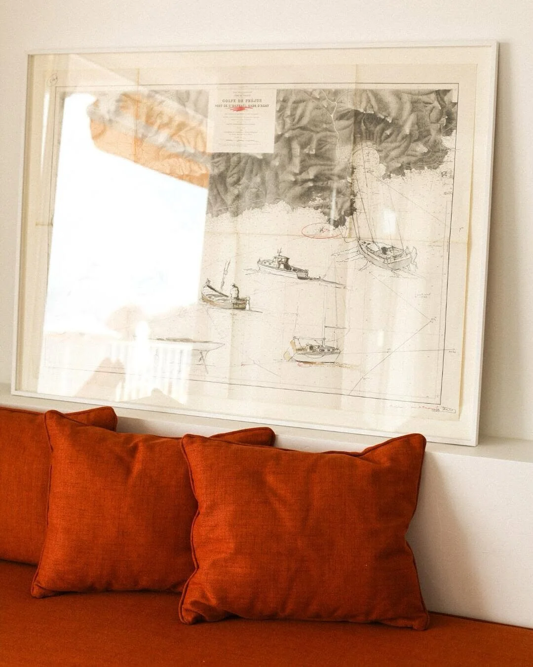 Framed nautical map with boats hanging above an orange couch with matching orange pillows.