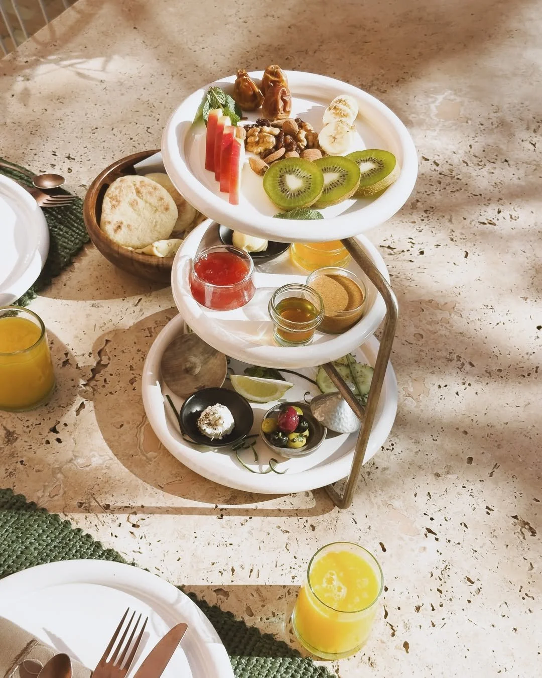 Tiered breakfast tray with sliced kiwi, watermelon, banana, dates, nuts, honey, jam, and cream cheese, with side dishes including bread, lemon wedges, and small bowls of syrup and butter, on a speckled beige table with orange juice and a fork and knife.