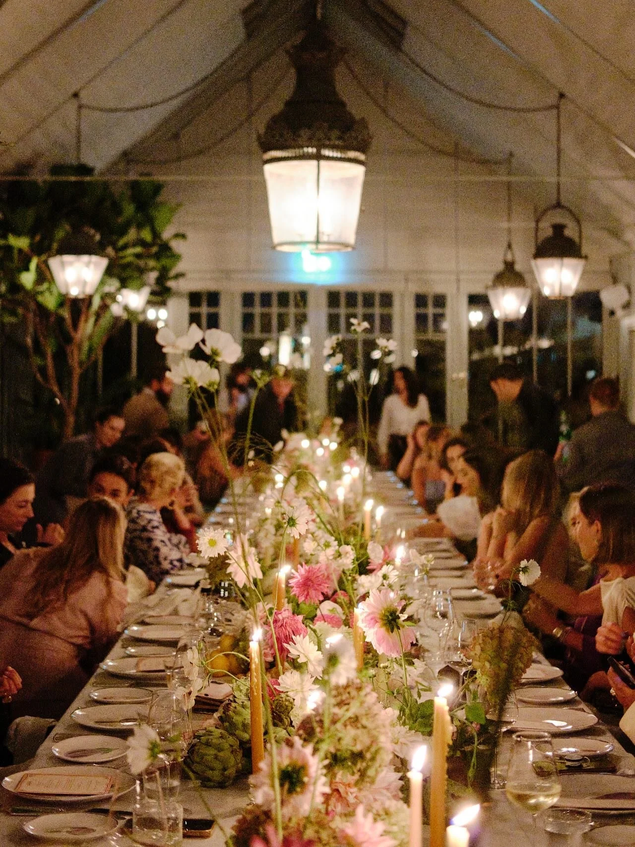 A long dinner table decorated with flowers and candles, surrounded by guests, in a warmly lit indoor space with large windows and hanging lantern-style lights.