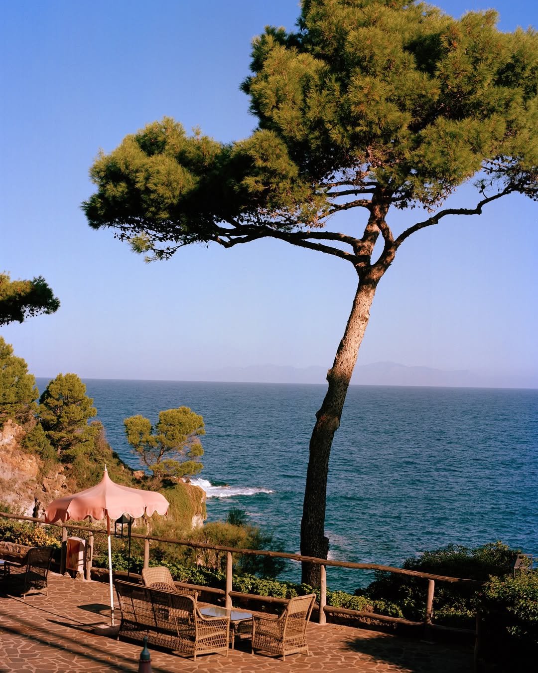Seaside patio with patio furniture and umbrella, tall leaning pine tree, overlooking ocean, clear sky, and distant mountains.