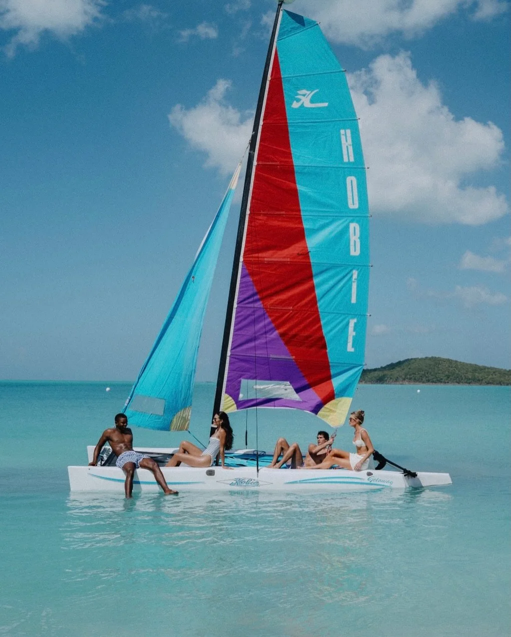 Four people relax and enjoy a sunny day on a colorful sailboat named 'Obie,' anchored in clear turquoise water with a small island or landmass in the background under a partly cloudy sky.