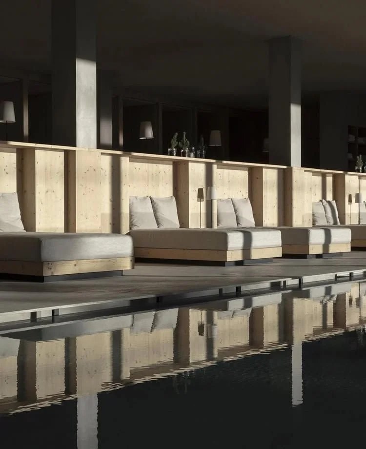Contemporary indoor lounge area with wooden panels, cushioned benches, and small lamps, reflected in a dark pool of water.