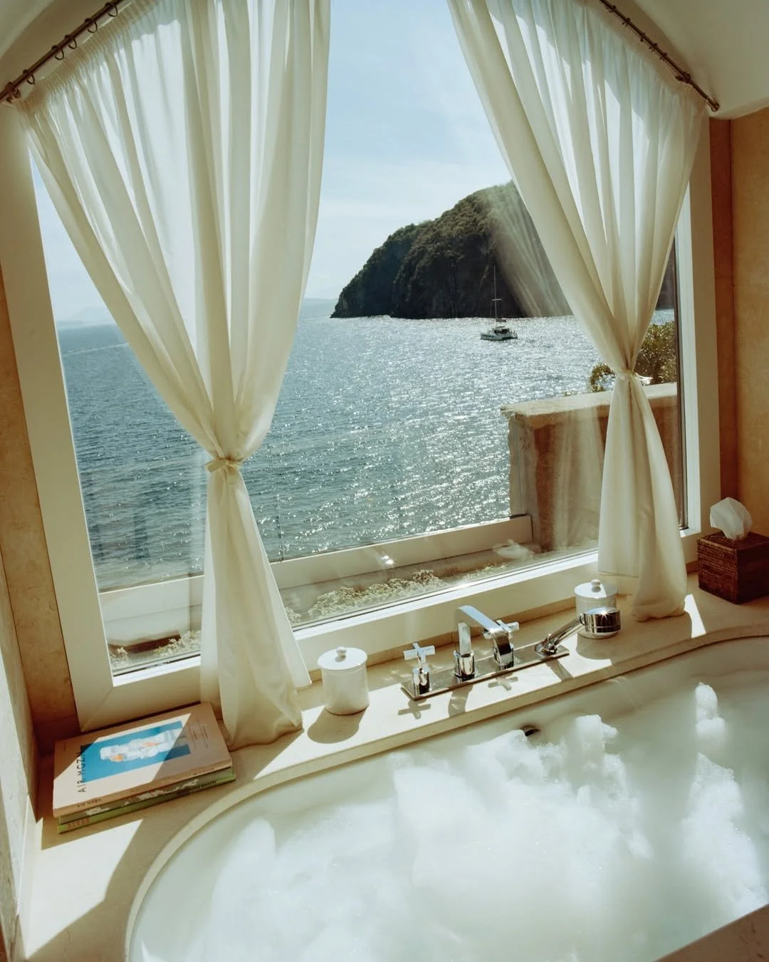A relaxing bathtub with bubbles in front of a large window overlooking the ocean and a rocky island with a sailboat, with cream-colored curtains and a decorative box on the side.
