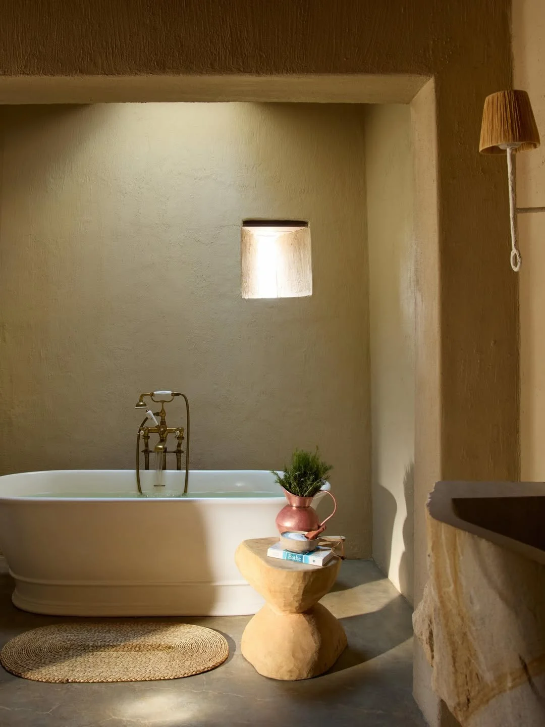 A bathroom with a white bathtub, a small window, a round woven rug, a wooden side table with a pink vase holding a green plant, and a bed of water. There is a small wall-mounted lamp on the right side.