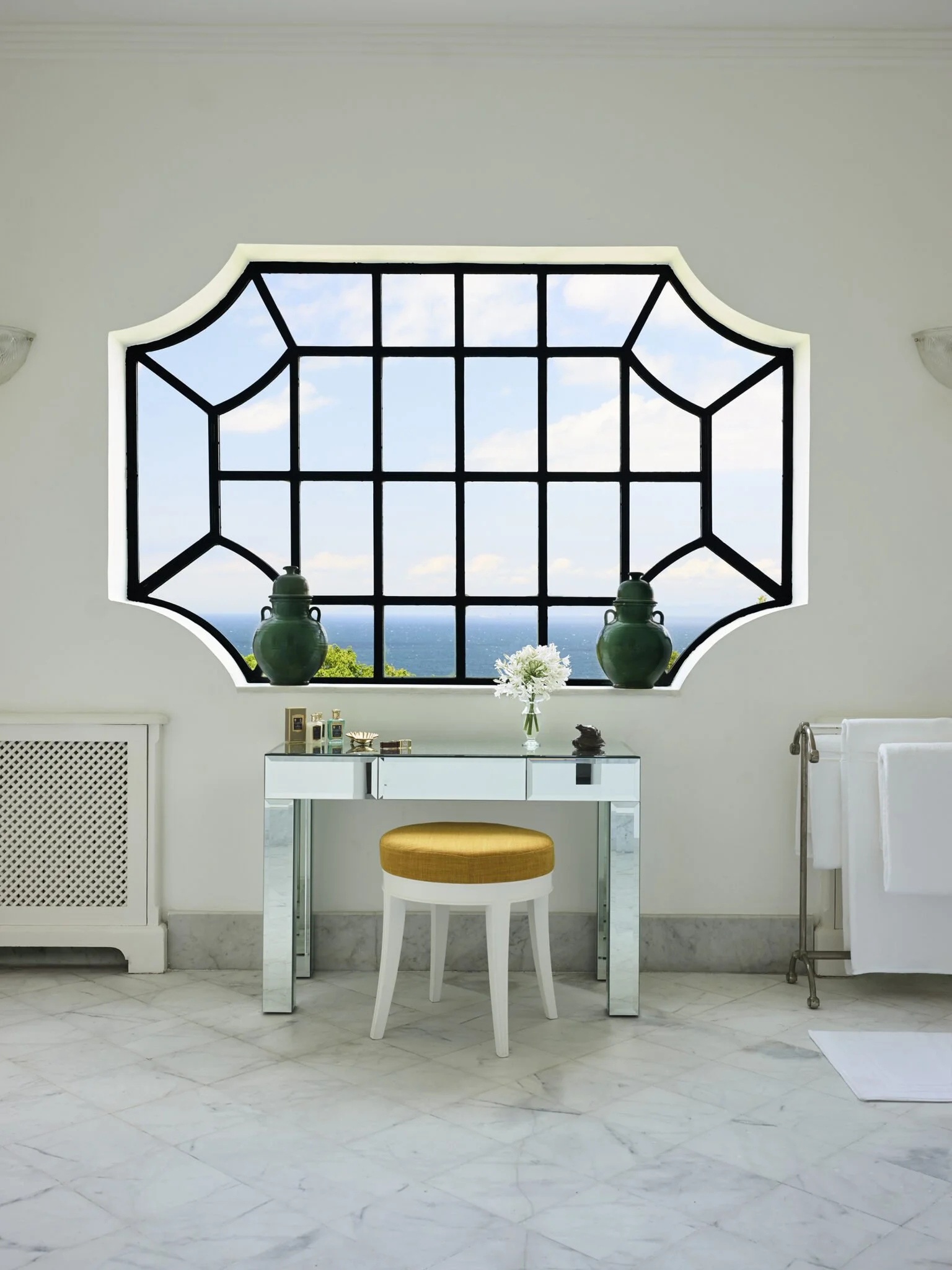 Interior view of a bright, white room with marble flooring and a large window with black framing, overlooking a view of the blue sky with some clouds. A small white desk with a mirror-like surface is centered in front of the window, with a round stool with a yellow cushion, a flower vase, and decorative items on it. To the right, a white towel hangs on a metallic towel rack, and on the left, a white radiator cover. Decorative lighting fixtures are mounted on the ceiling.