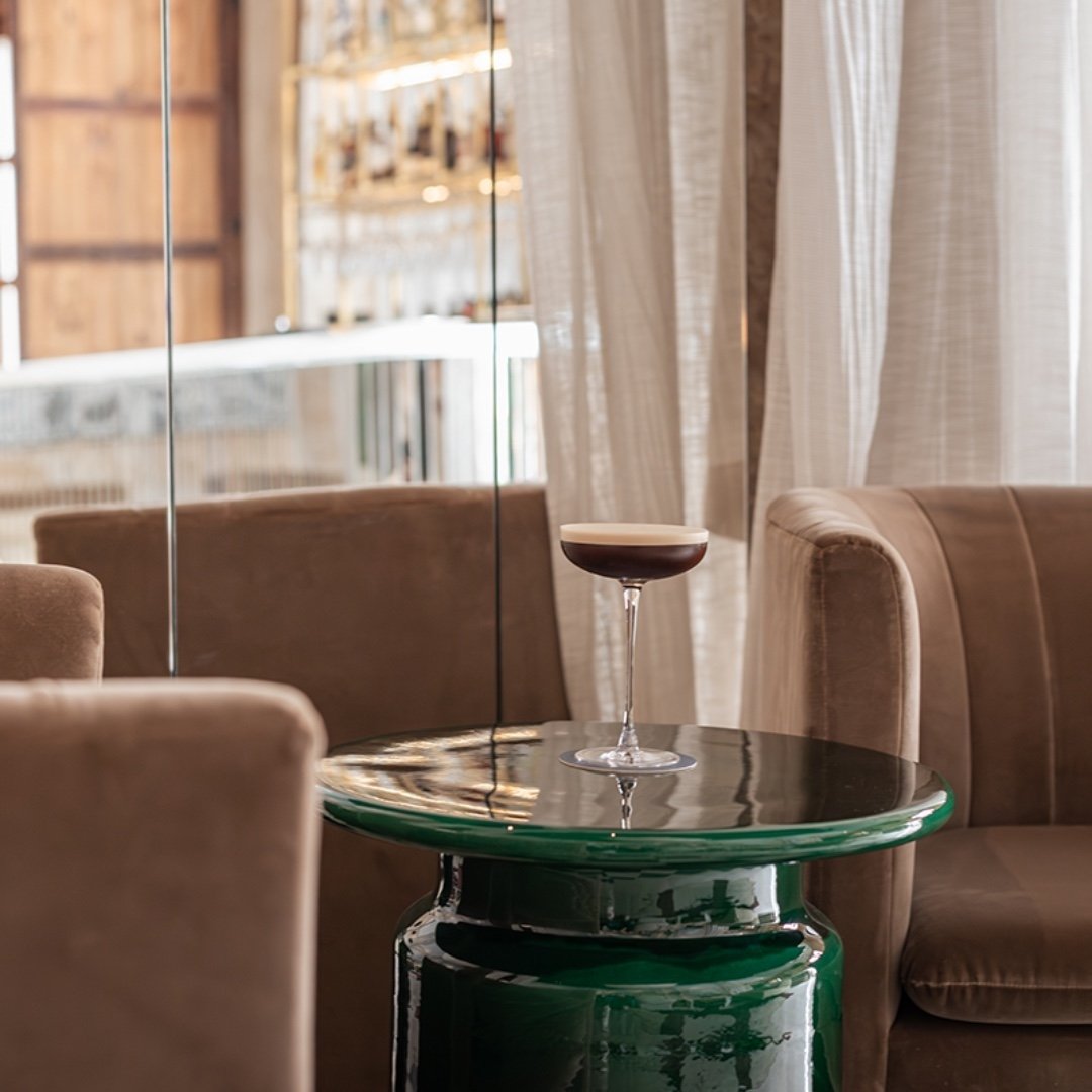 A cocktail glass with a dark beverage and a creamy foam top on a glossy green table, surrounded by beige upholstered chairs in a cozy, well-lit interior.