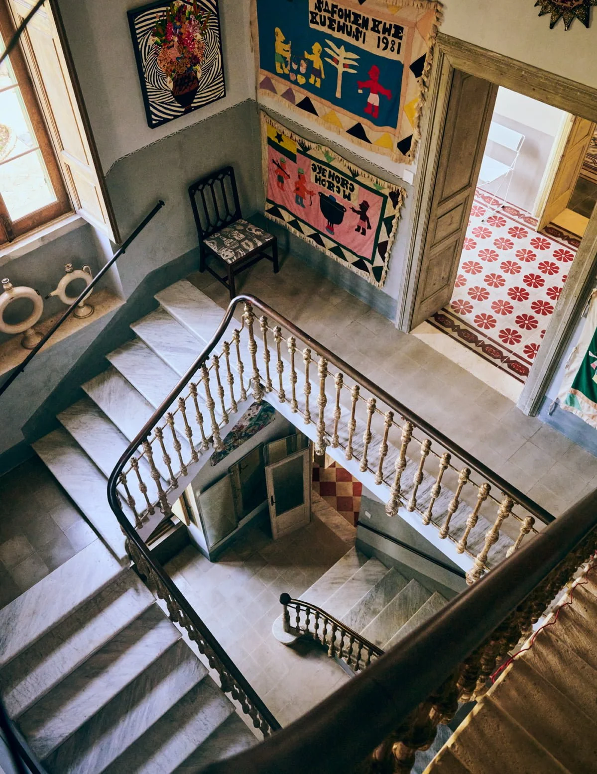 Interior view of a stairwell and upper hallway with colorful textile art on the walls, a black chair with patterned cushion, and a patterned tile floor outside an open doorway.