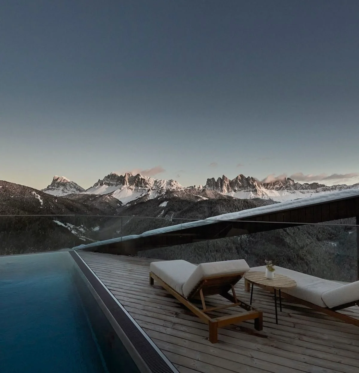 A mountain view from a wooden deck with two white lounge chairs and a small wooden side table holding a small plant. The deck overlooks snow-capped mountain peaks under a clear evening sky.