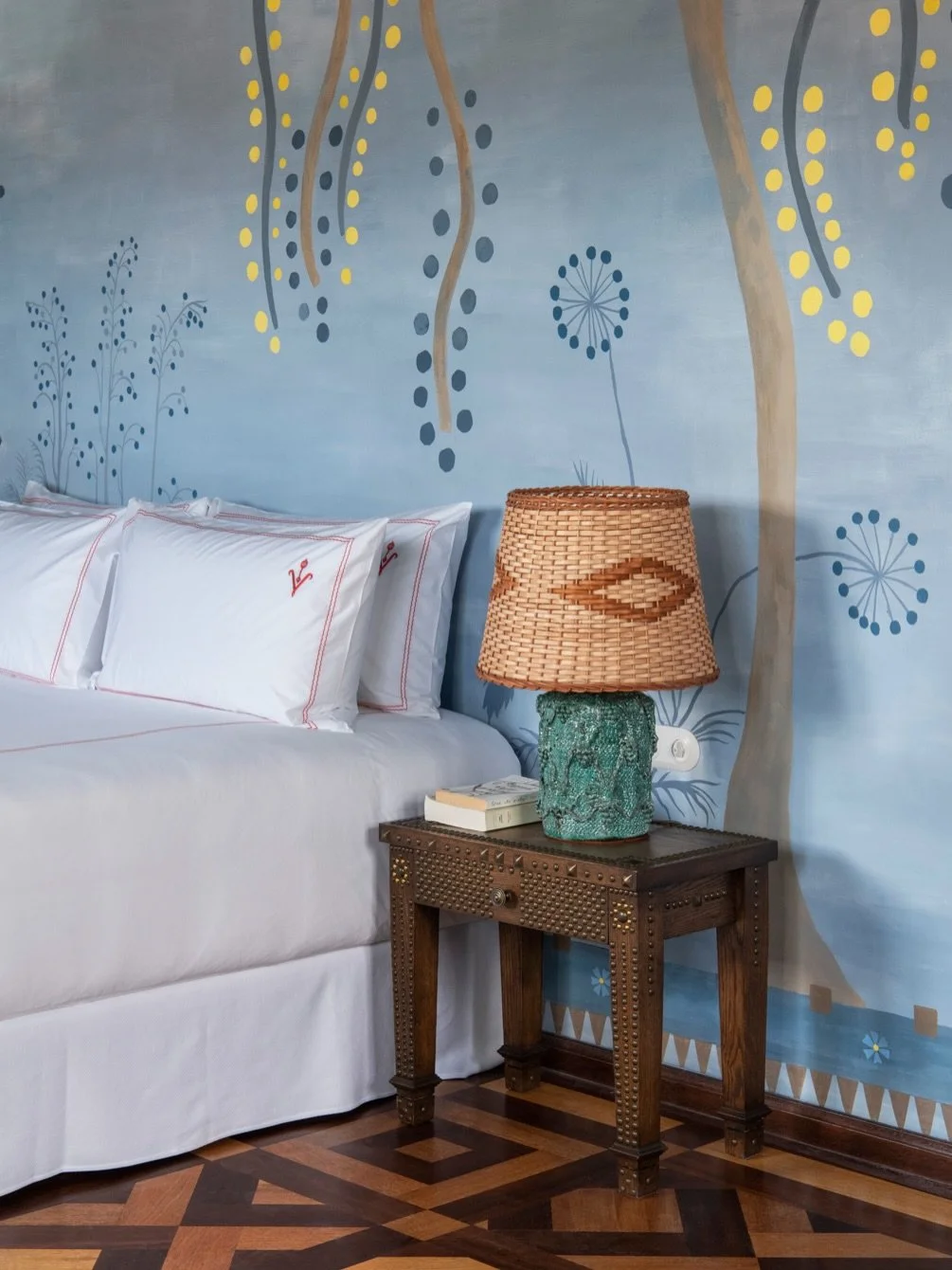 A bedroom with a white bed, decorative pillows, a wooden nightstand, a ceramic table lamp with a woven shade, books on the nightstand, and a blue wall with painted botanical and abstract designs.