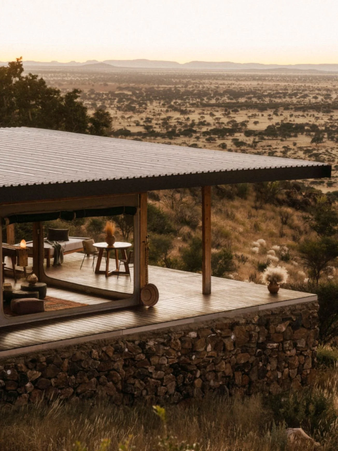 A small deck with outdoor furniture and potted plants, overlooking a vast desert landscape with sparse trees and distant mountains at sunset.