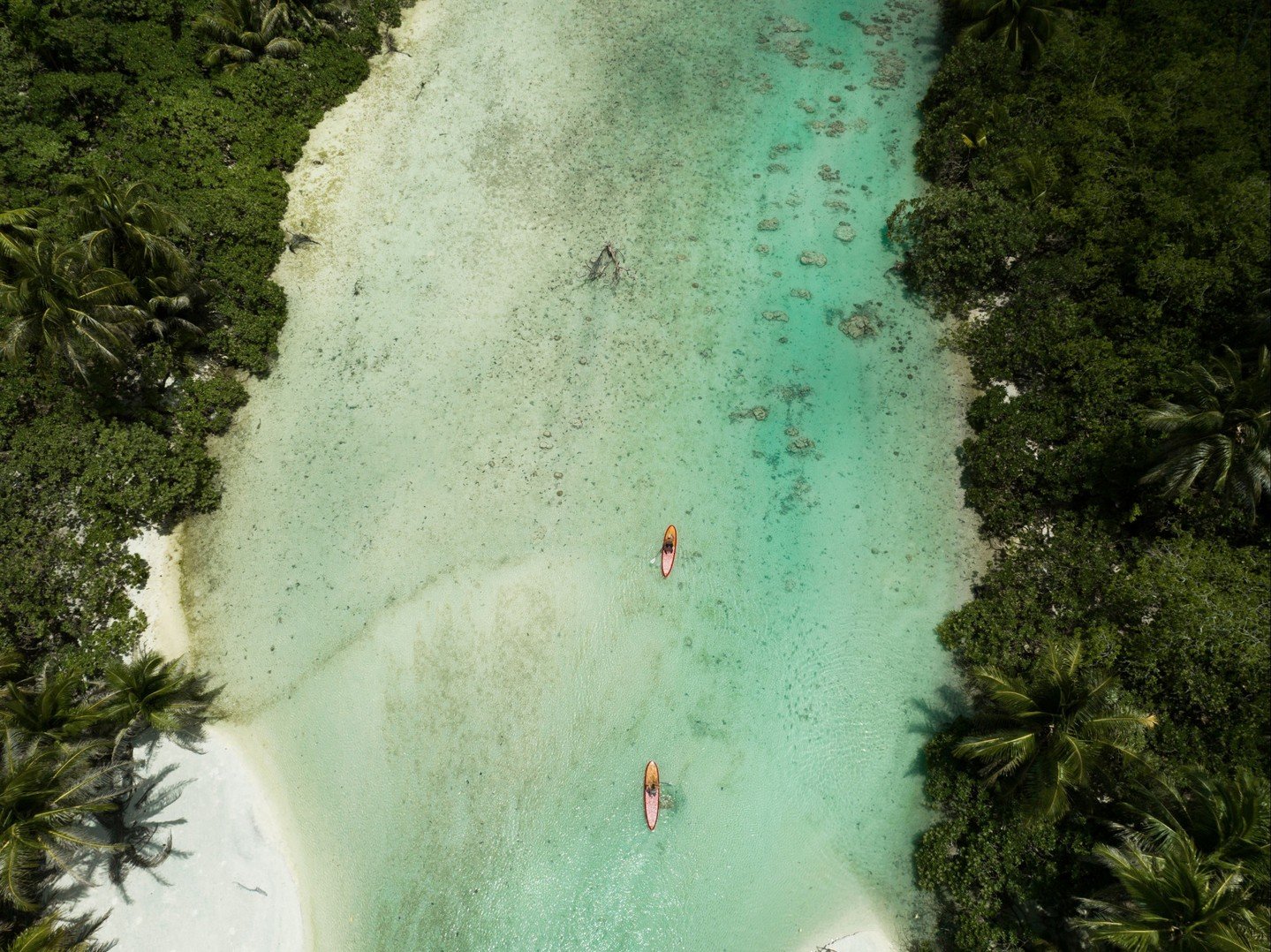 An aerial view of a narrow river with turquoise water, bordered by dense green tropical trees and foliage, with two kayakers paddling in the water.