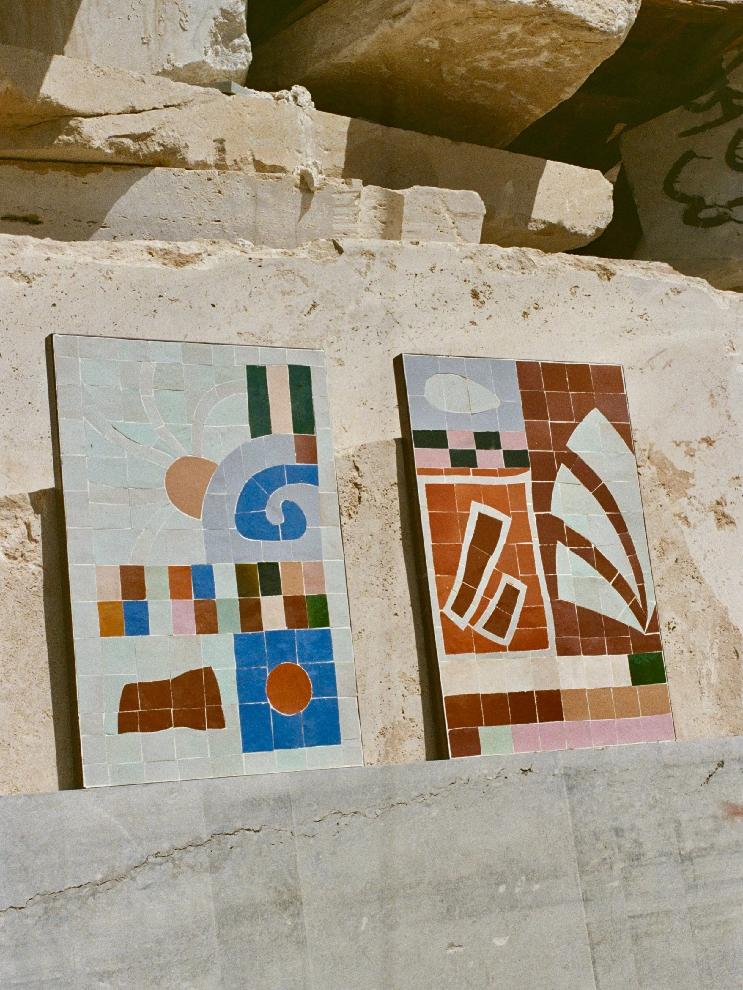 Two colorful mosaic art pieces with abstract geometric designs, leaning against a rough beige stone wall. One mosaic features a design with a large blue swirl, and the other has a pattern with white and red lines on a red background.