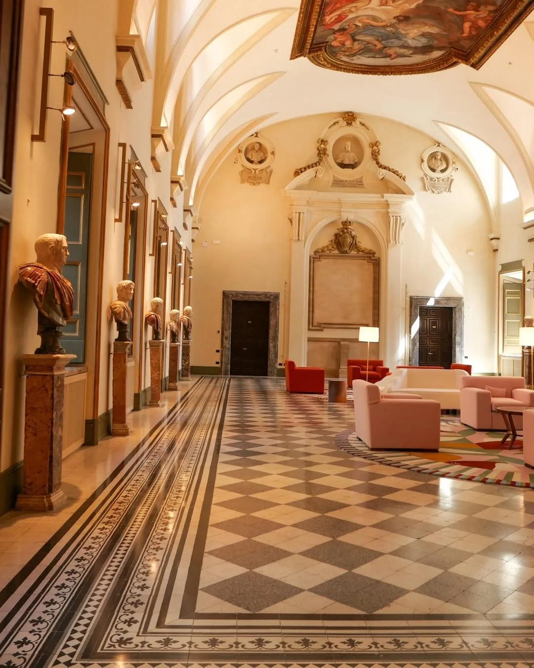 Elegant hotel lobby with bust sculptures along the left wall, high vaulted ceiling with ornate paintings, and a seating area with white and pink sofas and lamps.