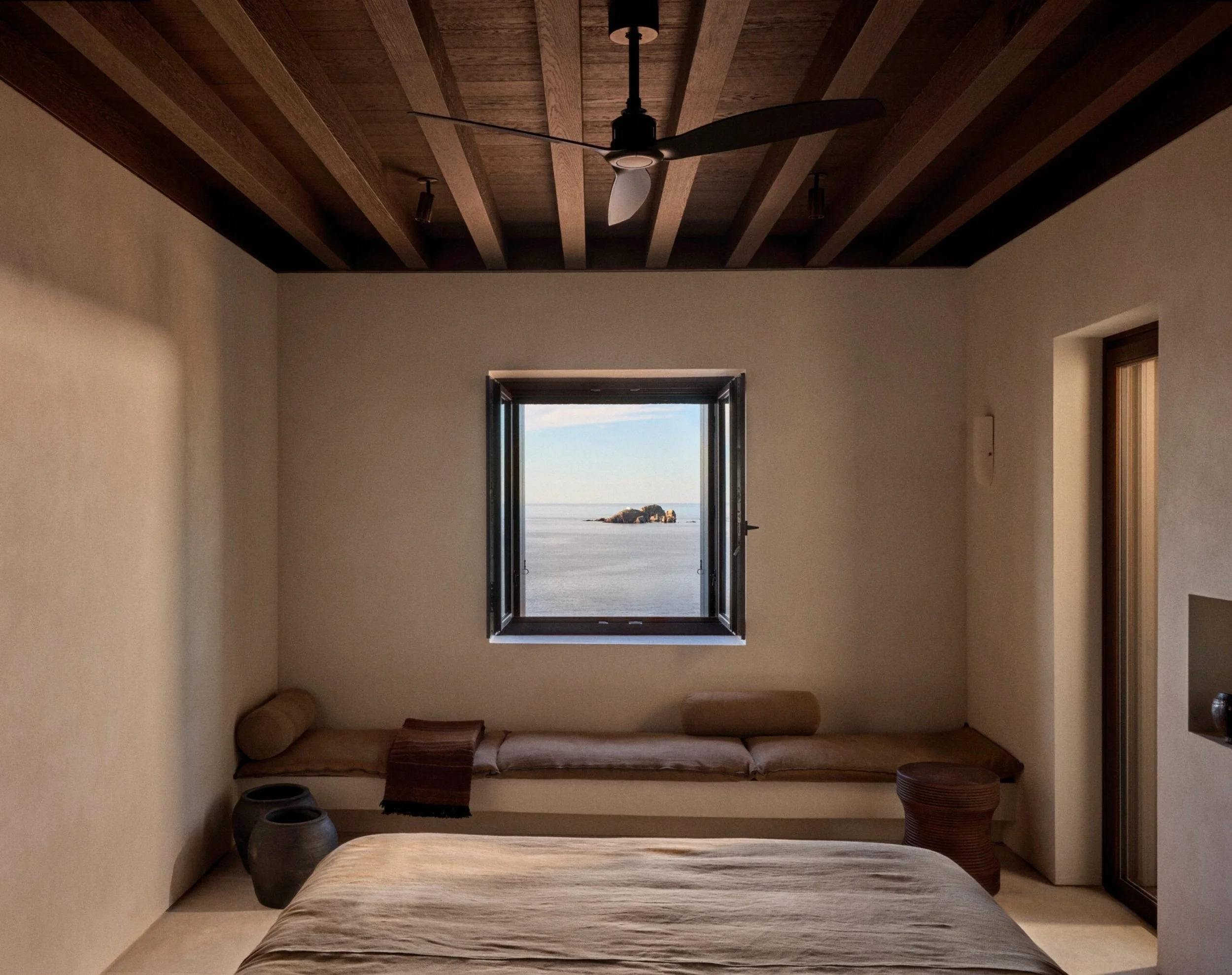 A cozy bedroom with a window view of the ocean and rocks. The room has beige walls, a wooden ceiling with beams, a ceiling fan, a bed, a cushion on the bed, and some decorative pottery.