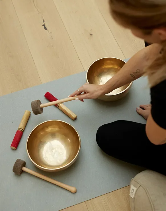 Person sitting on the floor with two singing bowls and mallets on a grey mat, preparing for sound healing session.