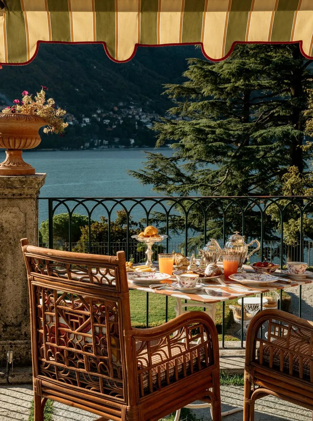 An outdoor breakfast table with floral china teapots, cups, glasses of orange juice, and plates of food, set on a terrace overlooking a lake and mountains, with trees and a decorative planter in the background.