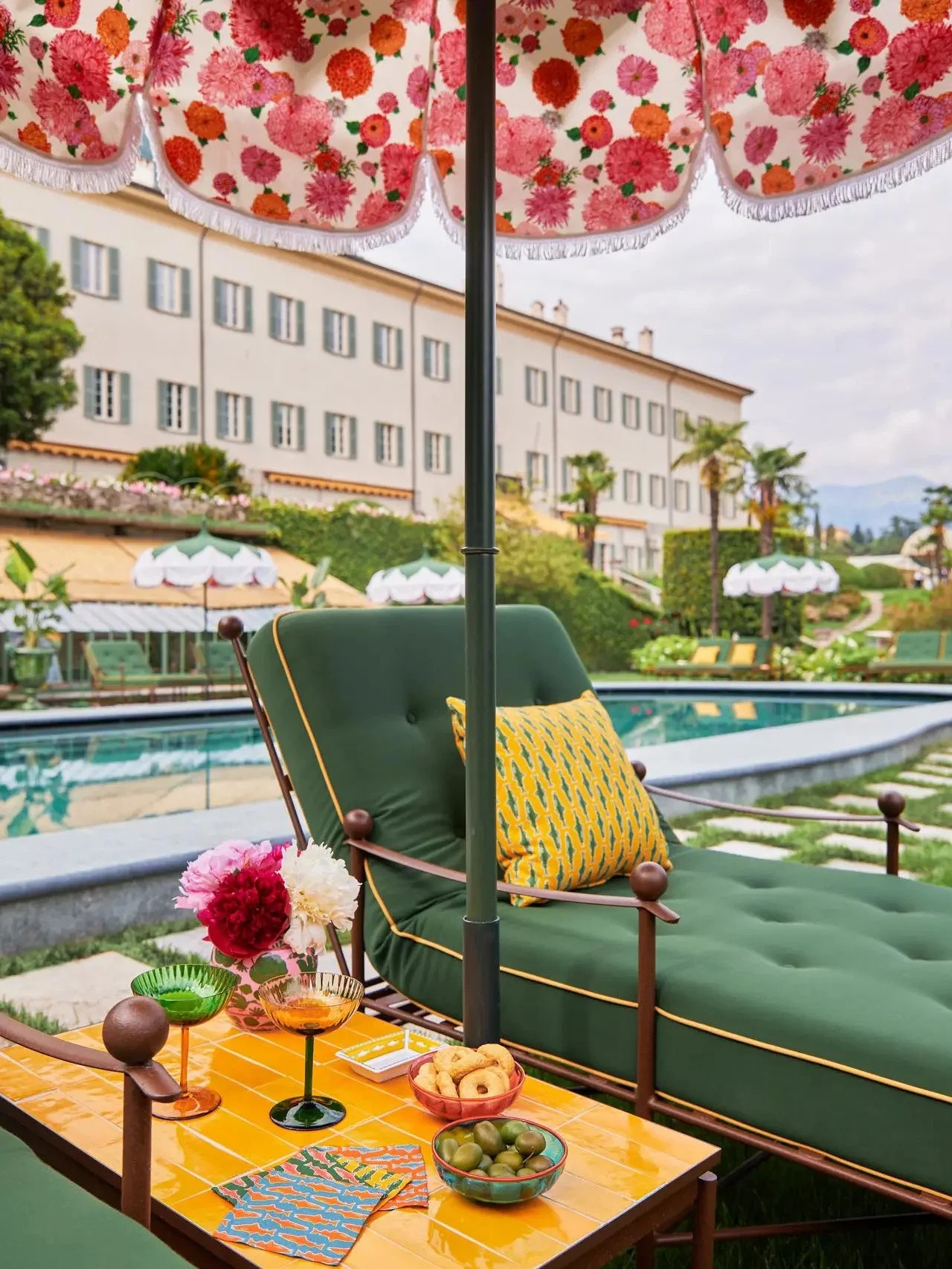 Picture of an outdoor poolside lounge area featuring a green cushioned chaise with a yellow patterned pillow, a floral umbrella, a tray with snacks and drinks, a pink and red flower arrangement, and a background of a large hotel building and palm trees.