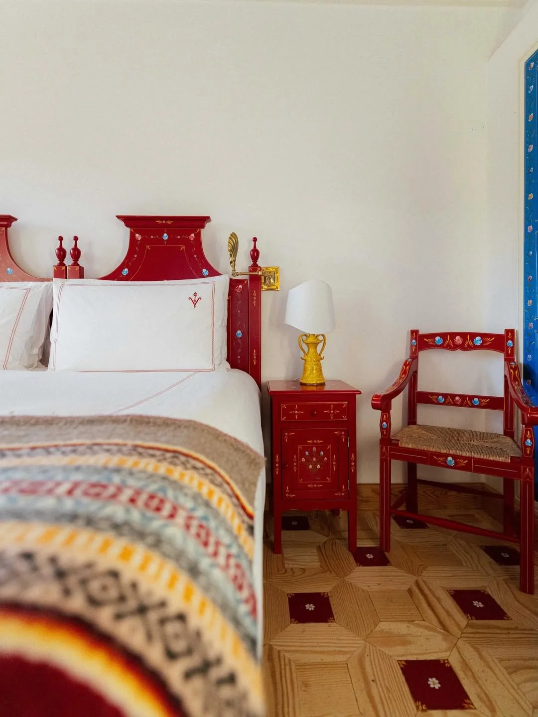 A bedroom with a white bed, red headboard and side tables, a yellow and white lamp, and a red wooden chair with floral designs on the headboard and armrests, and a hardwood floor with a geometric pattern.