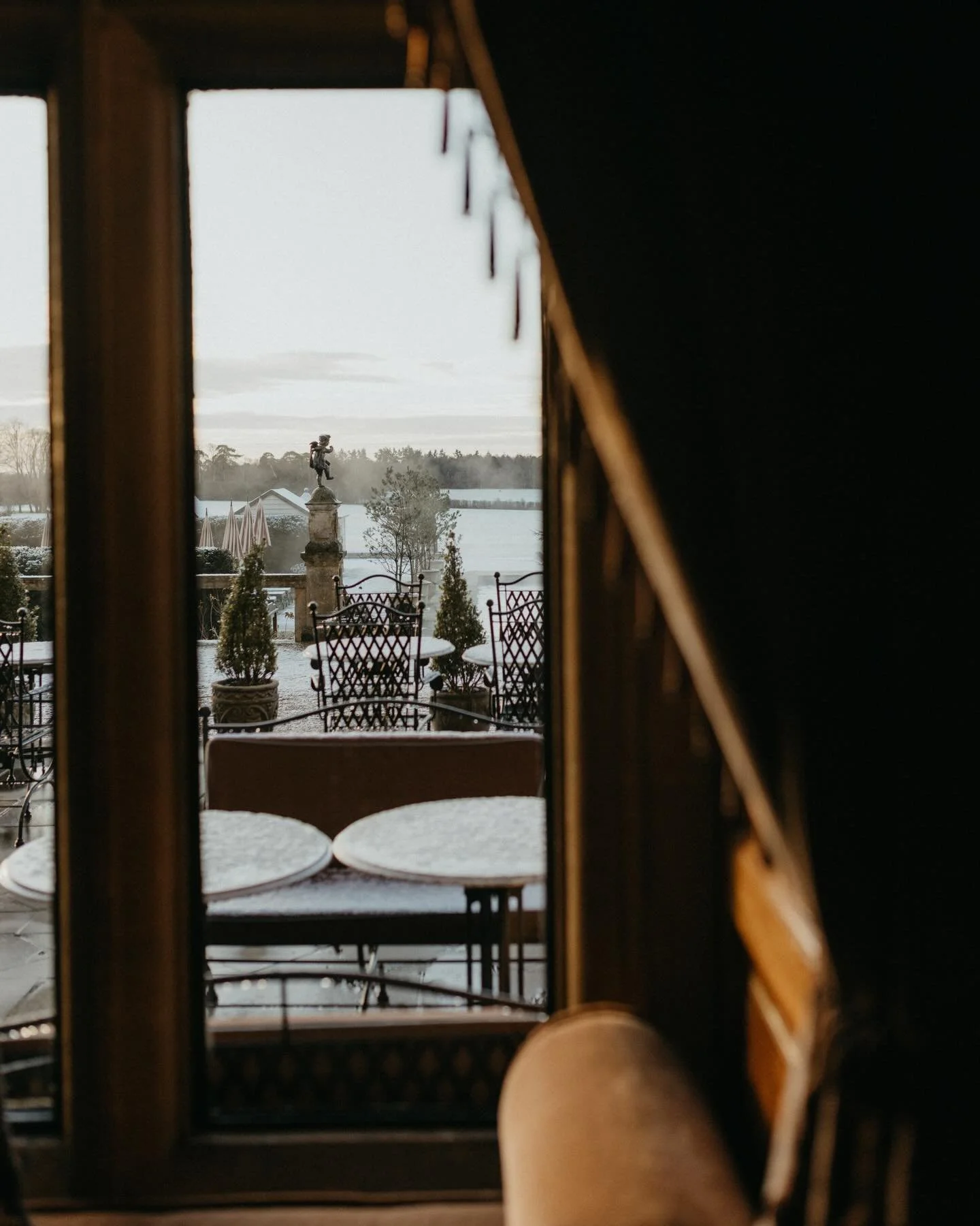 View of a snow-covered outdoor patio with metal chairs and potted plants, seen through a window or door frame, with a statue and a snowy landscape in the background.