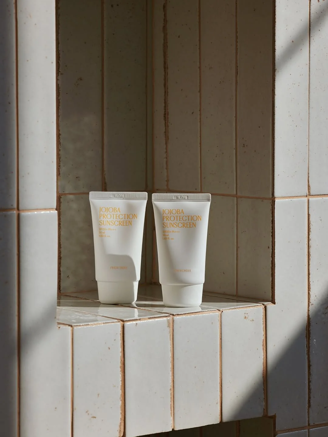 Two tubes of JoJoBA Protection Sunscreen placed on a tiled shelf with sunlight casting shadows.