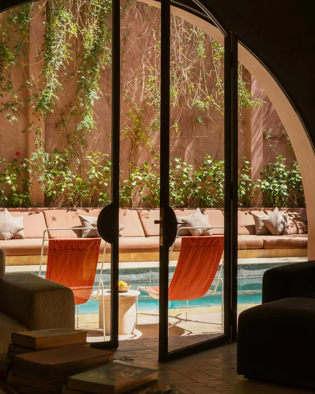Interior view of a room with a large glass sliding door leading to an outdoor pool area. Outside, there is a pink cushioned sofa, two orange chairs, a small side table with a bowl, and green climbing plants on a brown wall.