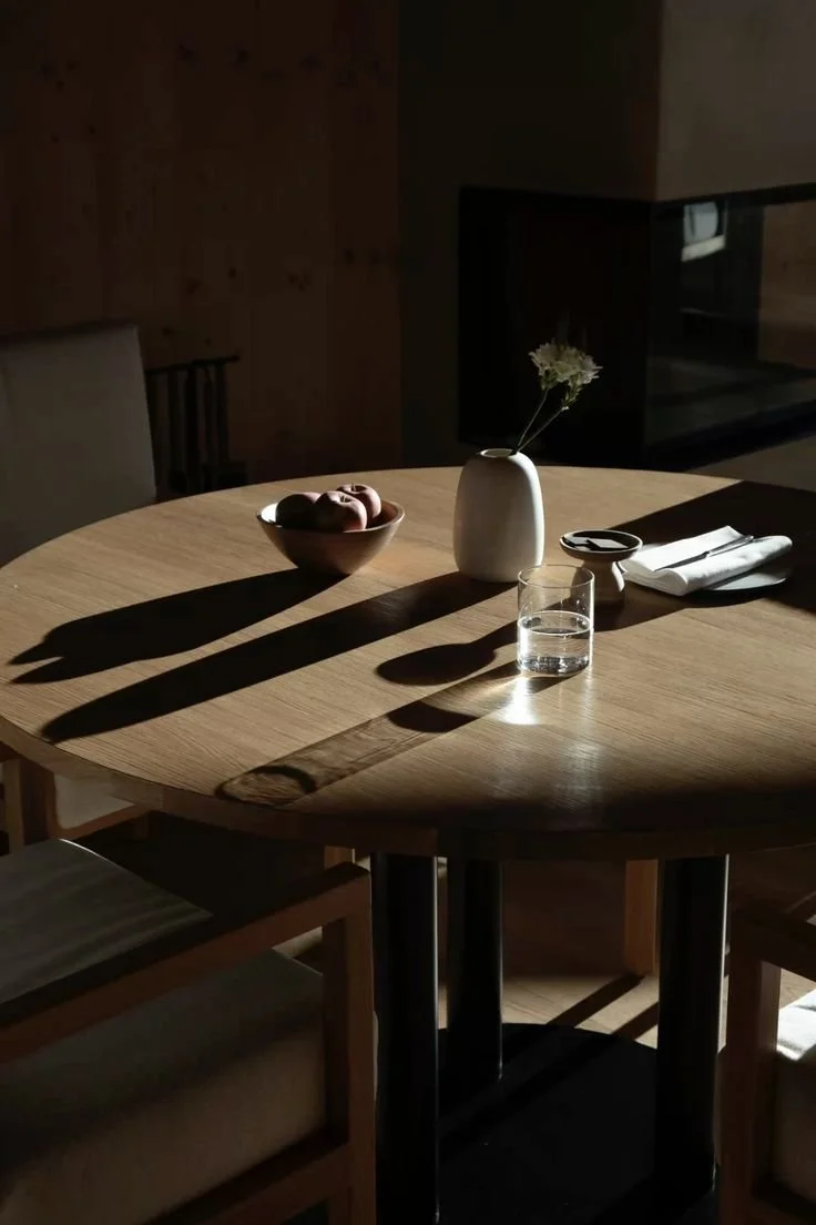A round wooden table with a bowl of fruit, a white vase with a flower, a glass of water, and a napkin with utensils inside, illuminated by sunlight creating shadows.