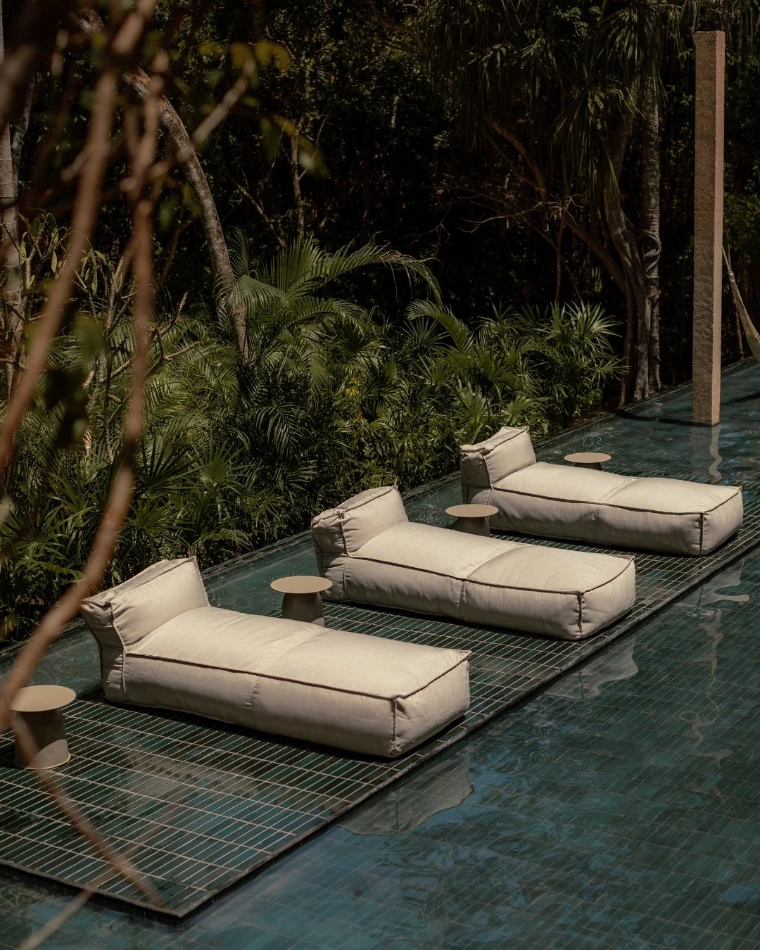 Three cushioned lounge chairs with small round side tables are placed on a tiled platform beside a pool, surrounded by lush green tropical plants and trees.