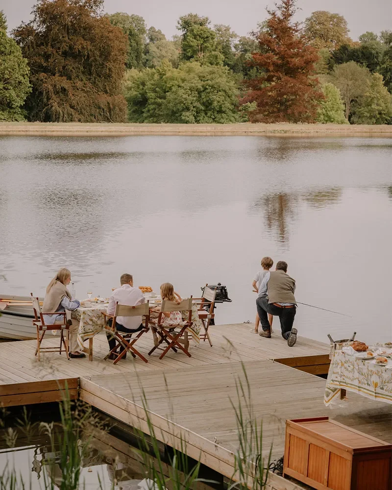 People enjoying a lakeside meal on a wooden deck, with two individuals fishing into the lake, surrounded by trees with autumn foliage.
