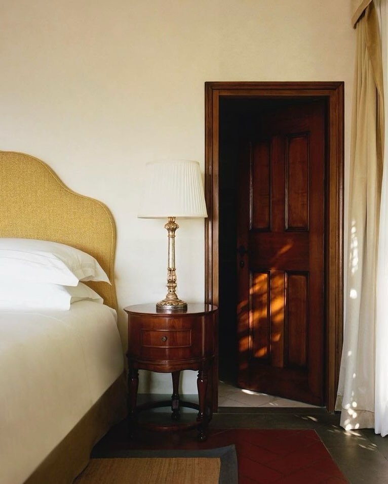 A bedroom nightstand with a lamp next to a bed and an open wooden door, with sunlight casting shadows on the door.