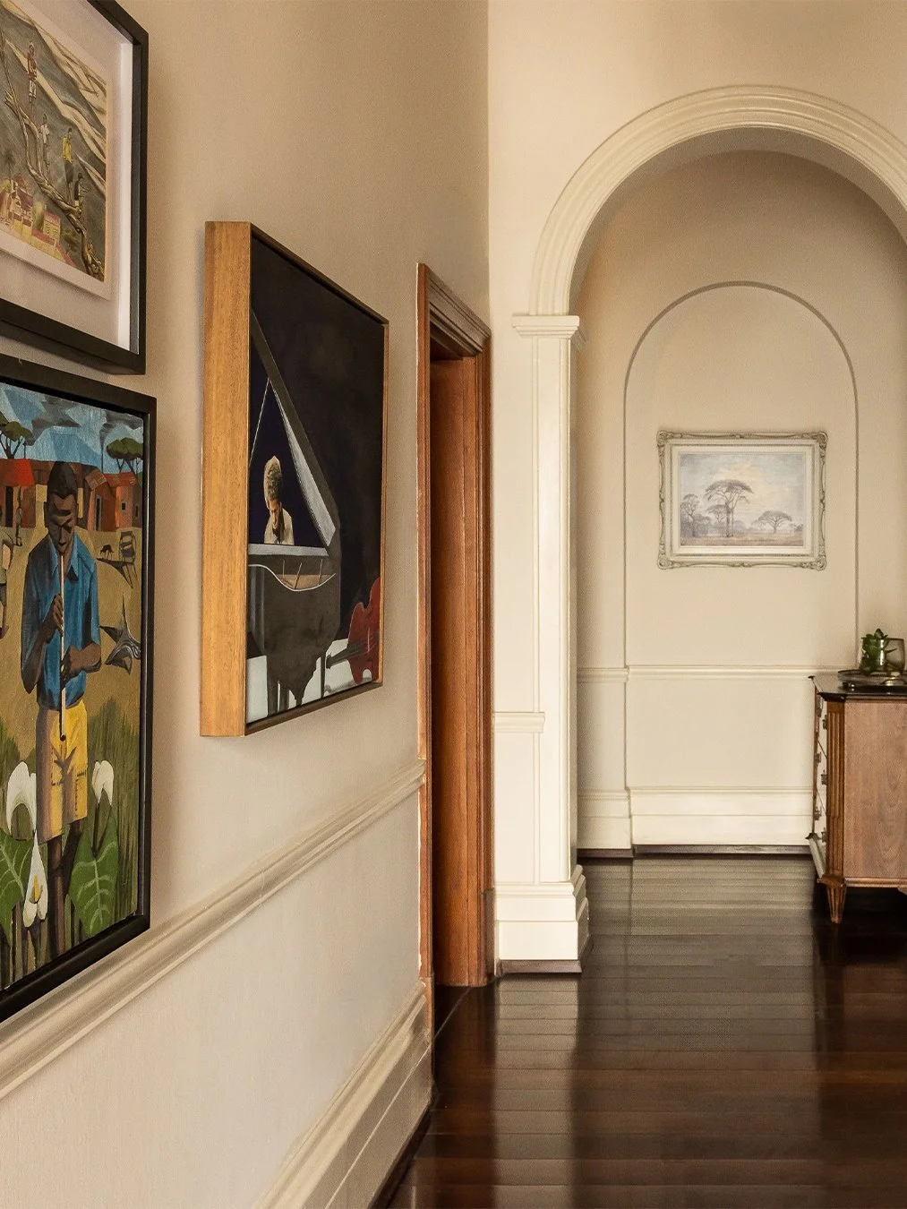 Hallway with framed artwork on the walls, arched doorway with decorative molding, and wooden furniture on dark wood flooring.