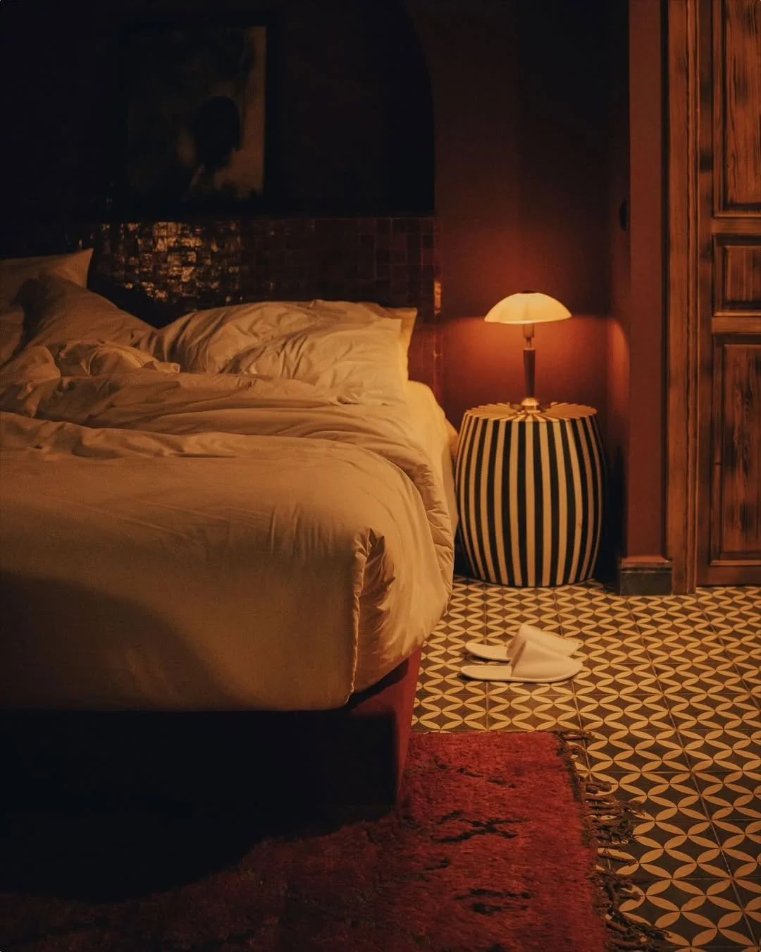 A cozy bedroom with a neatly made bed, a striped table lamp on a round side table, a pair of white slippers on the patterned tile floor, and a painting hanging on the wall above the bed.