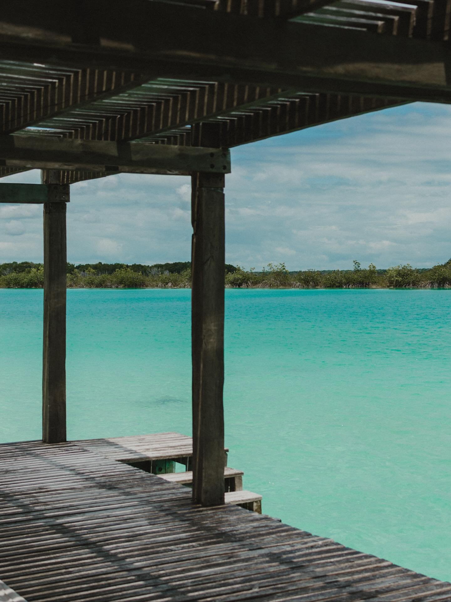 Wooden pier extending over turquoise water with a distant green shoreline and a partly cloudy sky.