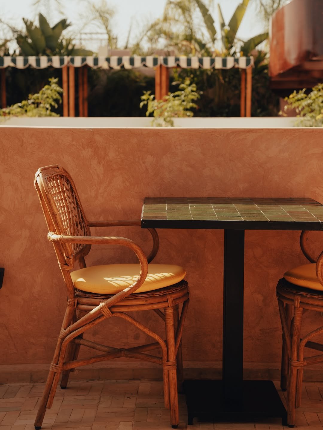 Wicker chairs with cushions and a small table set against an orange stucco wall, outdoor patio with greenery and striped awning in the background.