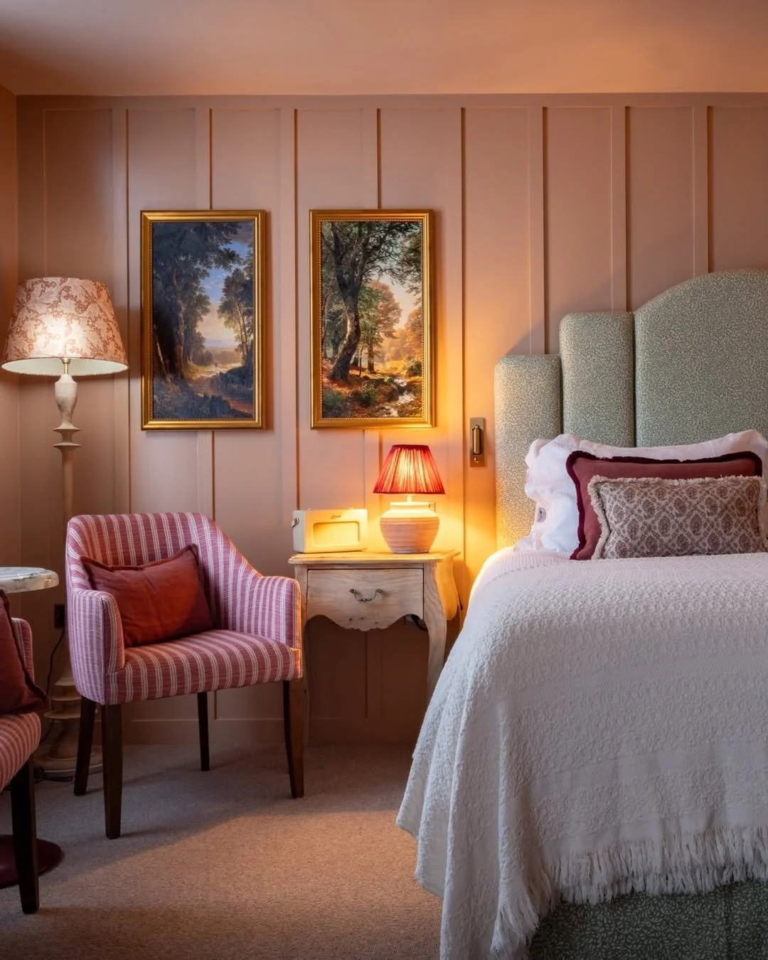 Cozy bedroom with a bed, pink and red pillows, a green upholstered headboard, two framed landscape paintings, a pink striped armchair with a pillow, a wooden nightstand with a red lamp, and a lamp with a floral shade.