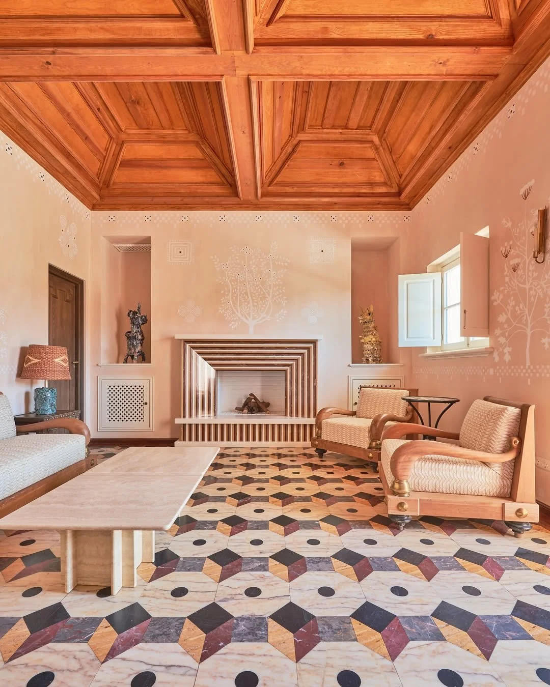 A living room with a patterned tile floor, pink walls with decorative white designs, a wooden ceiling, and furniture including two armchairs, a sofa, a coffee table, and decorative statues near a fireplace.