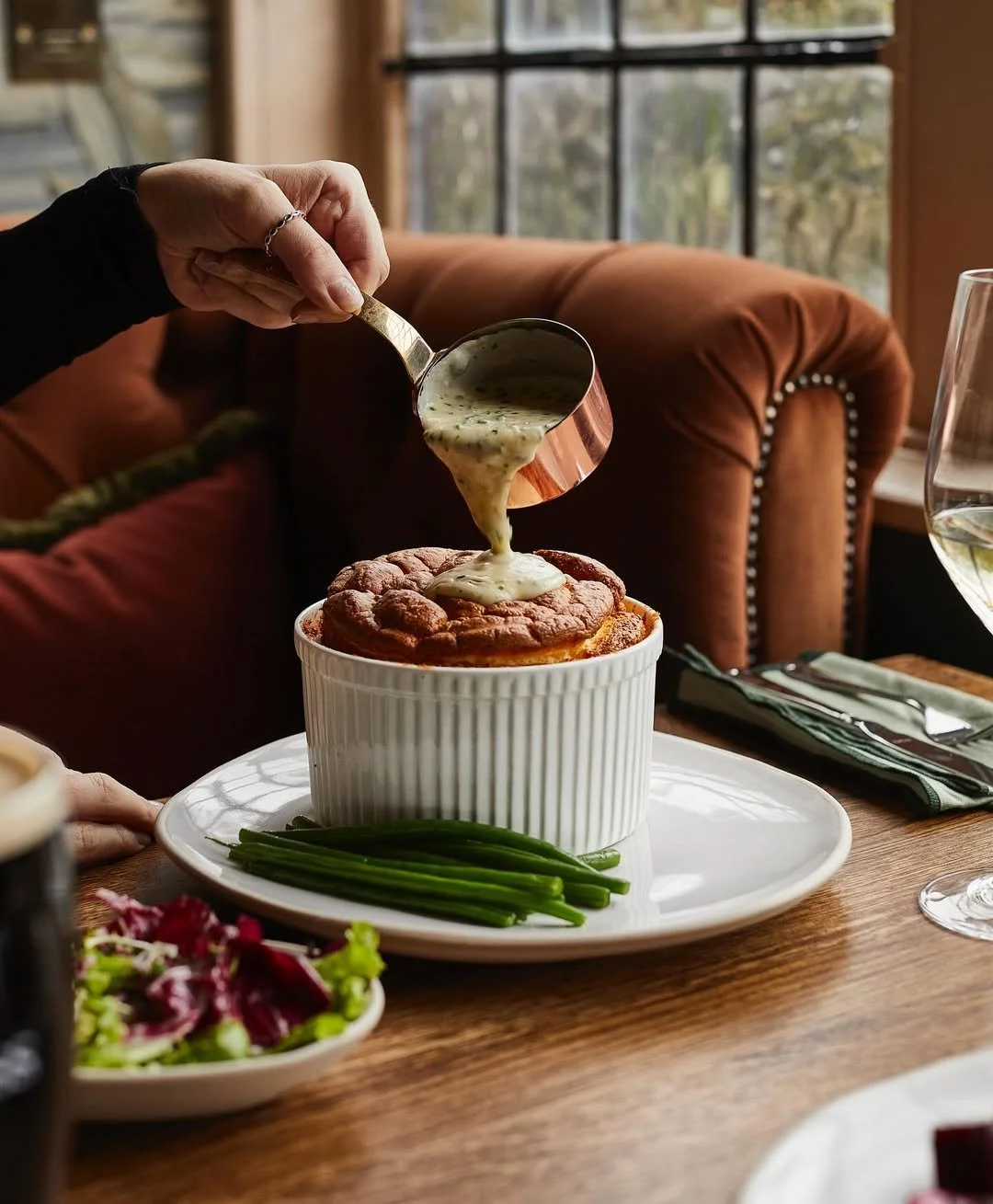 Person pouring creamy sauce over a sweet potato casserole topped with marshmallows, on a table with green beans, salad, and a glass of white wine.