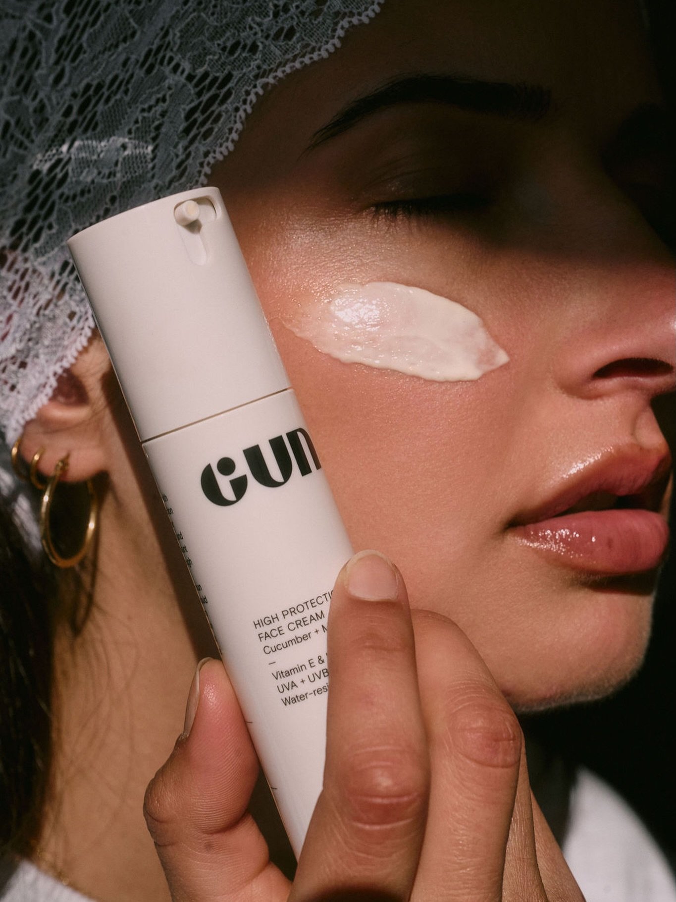 A woman with dark hair, earrings, and a lace head covering applies a white cream to her face with her fingers, holding a white tube labeled with skincare product information.