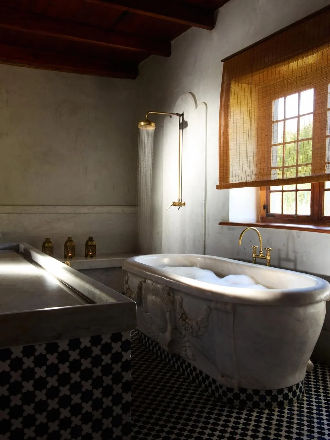 Elegant bathroom with marble clawfoot bathtub, vintage gold fixtures, window with wooden frame and bamboo shade, and a separate shower area with rainfall shower head.