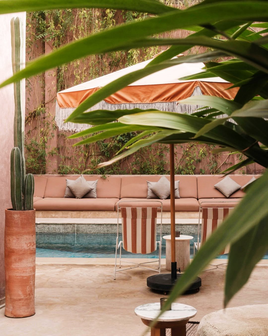 A stylish poolside area with tan outdoor sofa, striped chairs, large pink and white umbrella, cactus in a terracotta pot, and lush green plants and vines on the wall.
