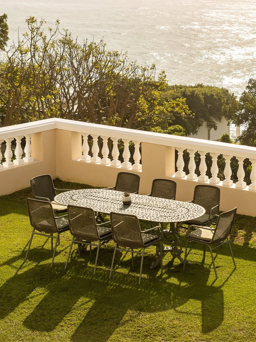 Outdoor patio with a white ornate table and eight chairs on a grassy area, overlooking trees and water in the background.