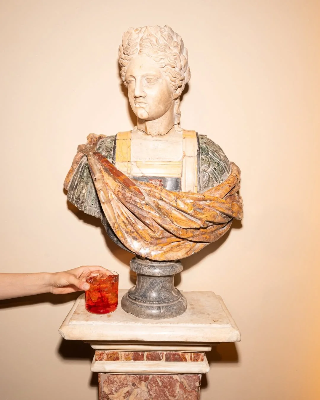 A classical marble bust sculpture of a woman with curly hair, draped in a colorful garment, on a pedestal. A hand on the left holding a glass with a red liquid and ice cubes is partially visible.
