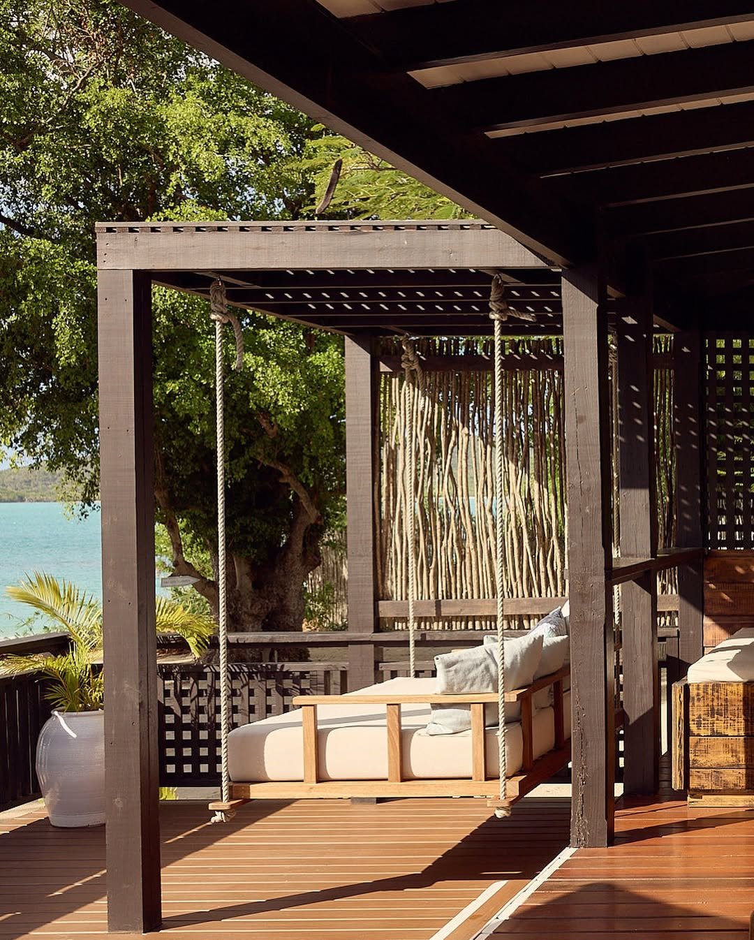A wooden outdoor deck with a white cushioned swing hanging from the ceiling, surrounded by lush green trees and overlooking a body of water.
