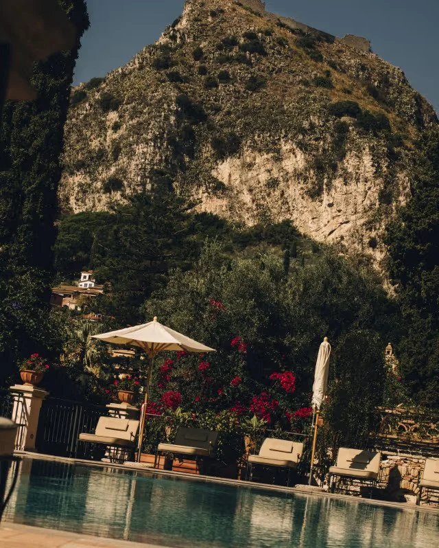 Poolside seating area with umbrellas and chairs in front of a lush garden with red flowers, backdrop of a large rocky mountain under a clear sky.
