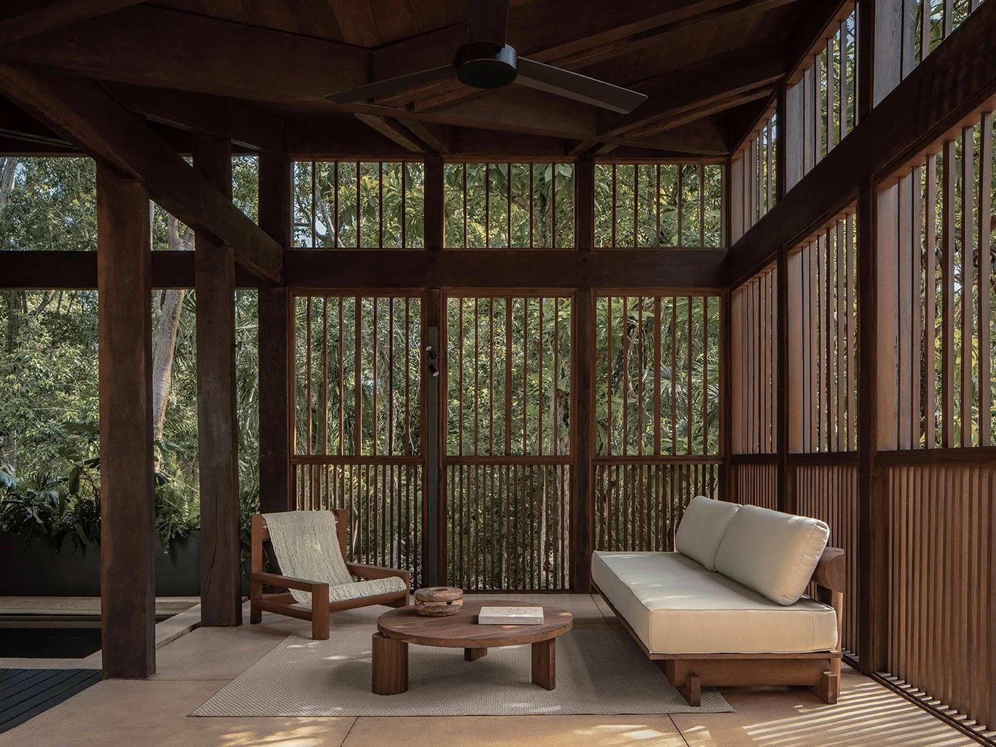 A screened porch with wooden framing, outdoor furniture, and view of lush trees outside.