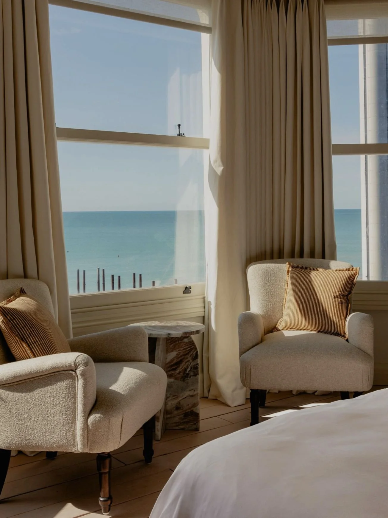 A cozy hotel room with beige armchairs, striped pillows, a small marble side table, and large windows with cream curtains showing a view of the ocean.