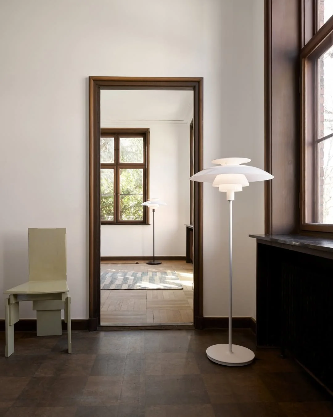 Interior view of a room with hardwood floors, a white wall, and wooden window frames. There is a small white chair to the left, a floor lamp with a white lampshade near the right wall, and another lamp with a larger white shade further inside the adjacent room, visible through a doorway.