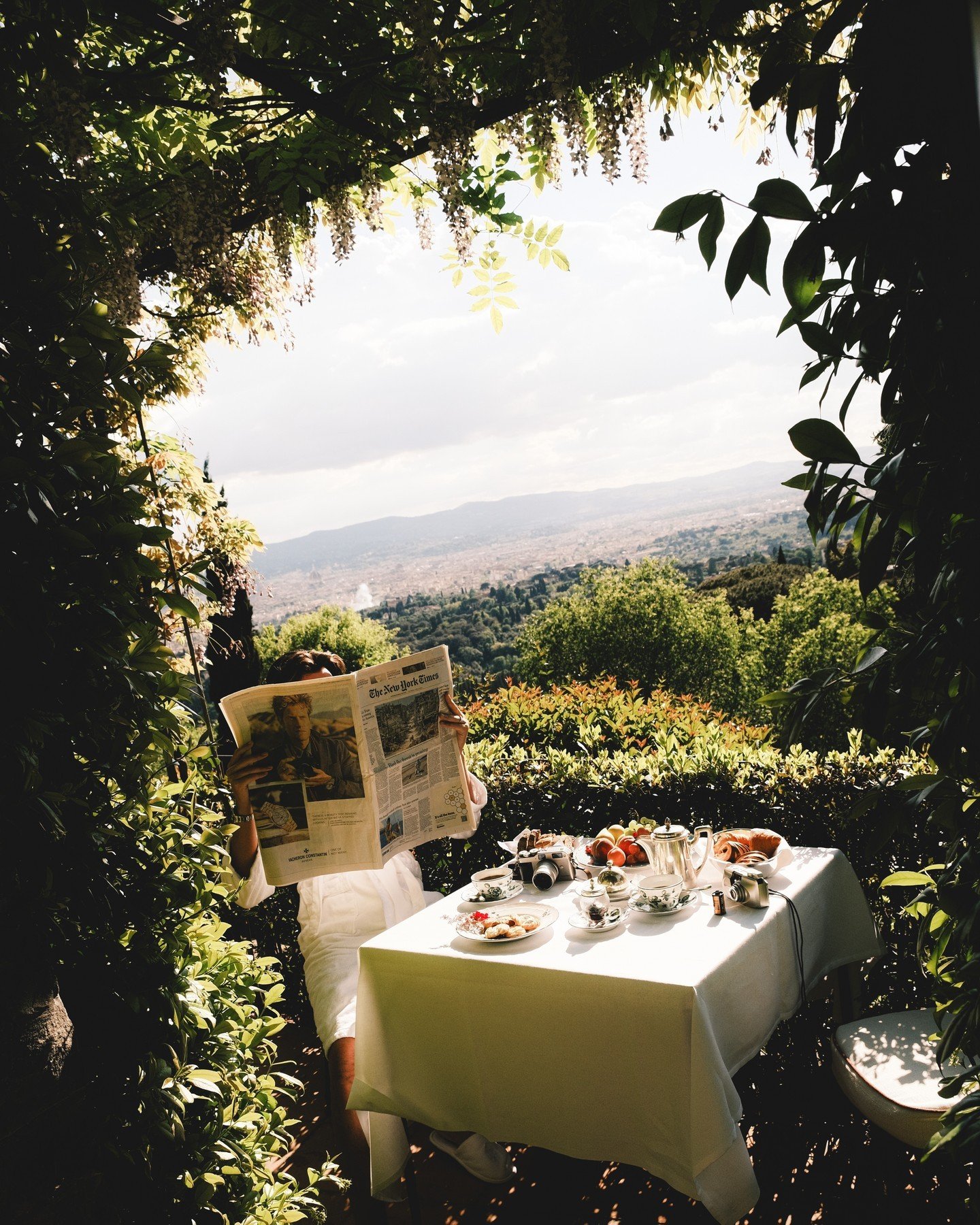 Person sitting outdoors at a table with a white tablecloth, drinking tea and reading a newspaper, surrounded by lush greenery and trees, with a scenic view of a cloudy sky and distant mountains.