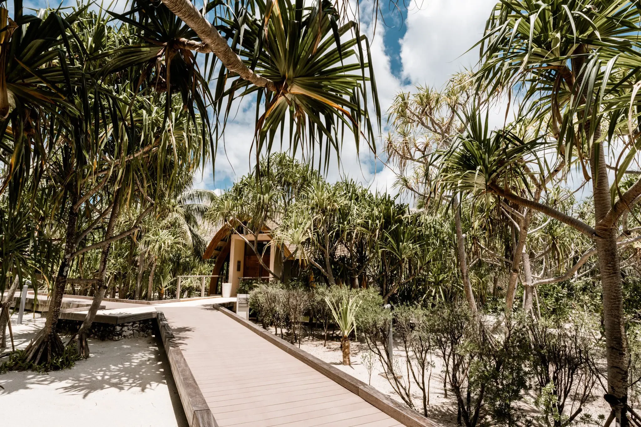 Pathway through tropical trees, with a small house visible in the background beneath a partly cloudy sky.