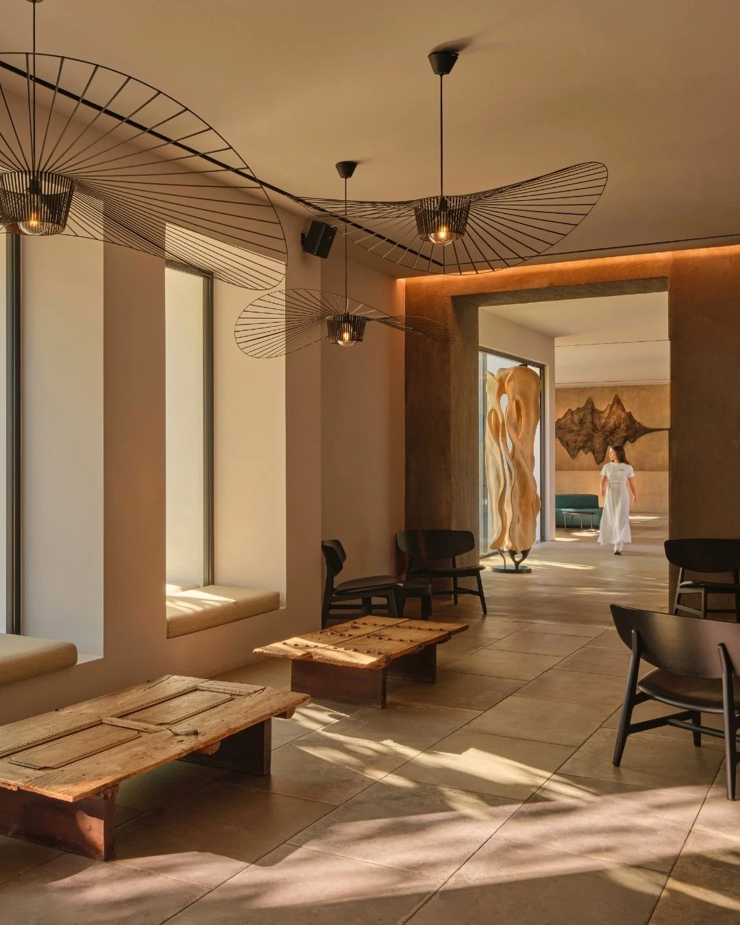 Modern interior space with sunlight coming through tall windows, wooden tables, black chairs, artistic ceiling light fixtures, a large wooden sculpture, and a woman walking in the background.